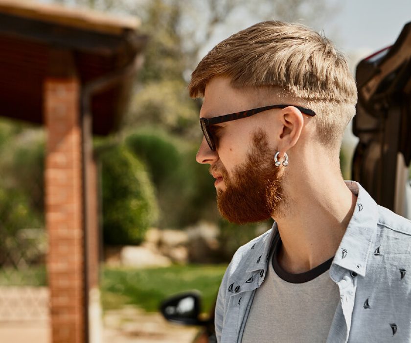 Un homme barbu et portant des lunettes se tient à côté d'une voiture, illustrant les tendances de mode masculine pour 2026.